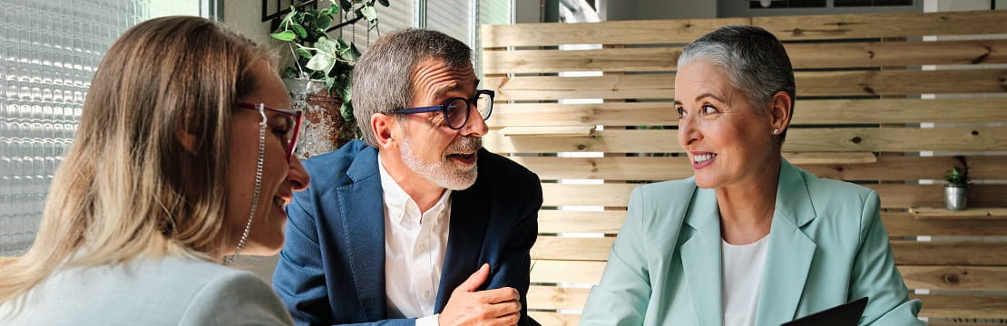 Three adults are speaking to one another and are seated in a warm, contemporary office space.